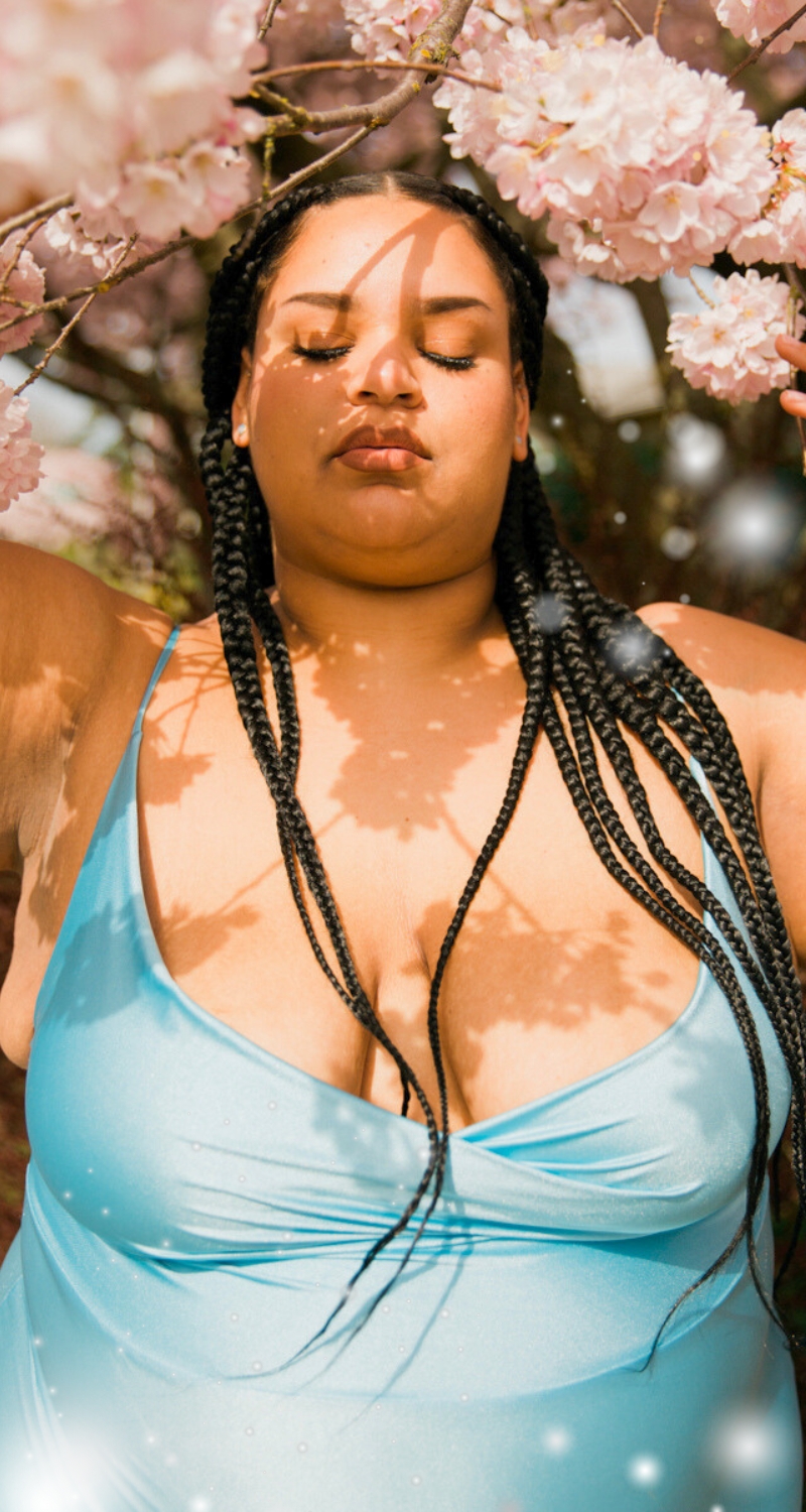 a fat positive plus sized mixed race light skinned pregnant woman with foliage and pink flowers behind her has her eyes closed with a peaceful look on her face and shadows from the branches on her face, she has long braided black hair and a light blue spaghetti strap silky dress on